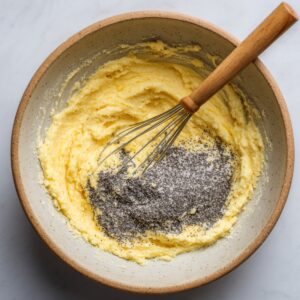 A ceramic bowl with pale yellow creamed butter and sugar, topped with poppy seeds, and a whisk resting inside.