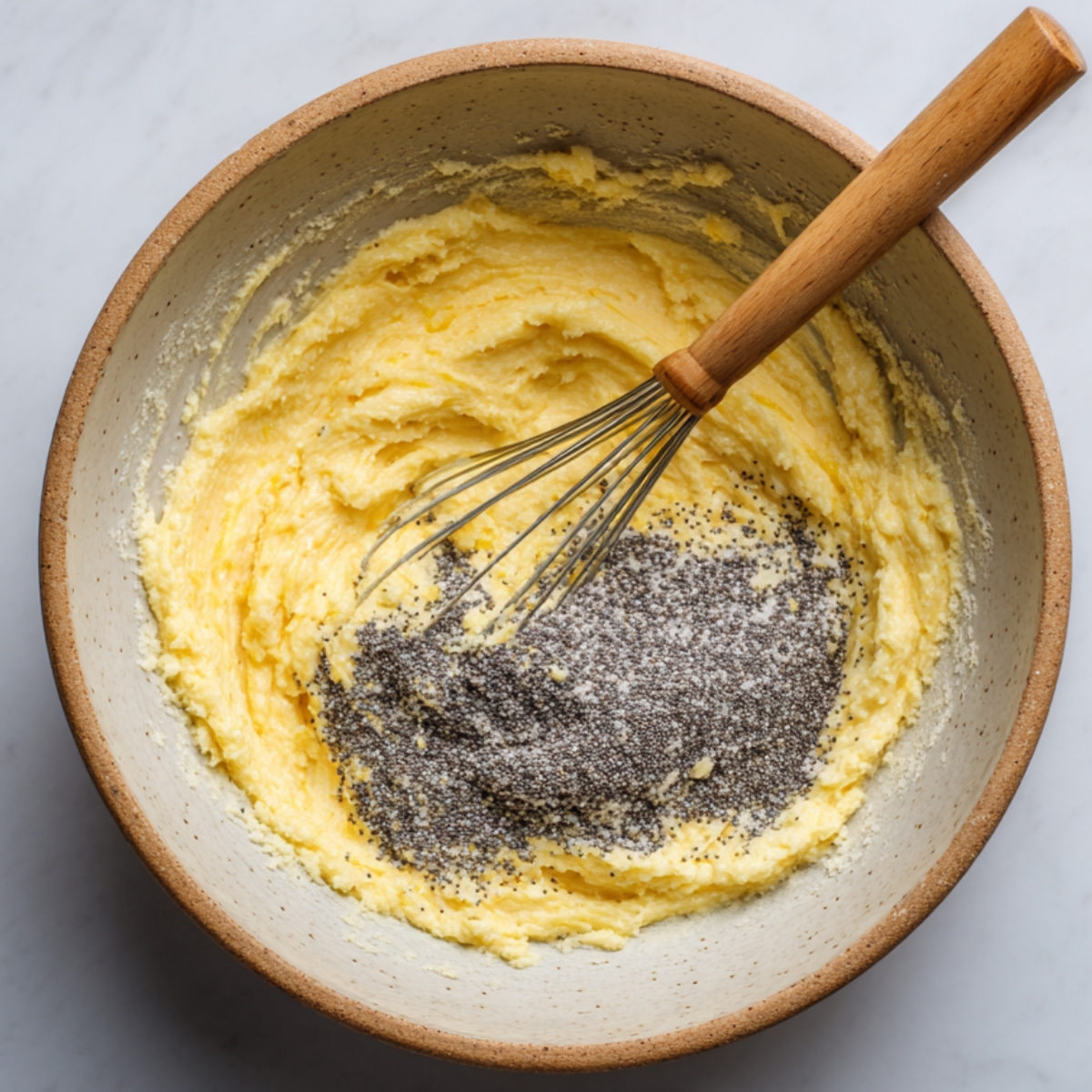 A ceramic bowl with pale yellow creamed butter and sugar, topped with poppy seeds, and a whisk resting inside.