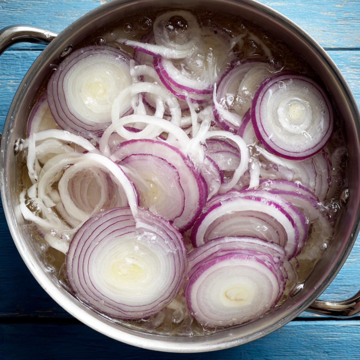 Sliced red and white onions soaking in ice water inside a stainless steel pot, on a bright blue wooden surface.