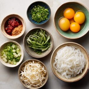 Overhead view of Pad Thai ingredients in small bowls, including eggs, rice noodles, scallions, chili paste, cilantro, peanuts, bean sprouts, and julienned green vegetables.