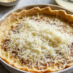 Delicious Parmesan Onion Squares Recipe 20 Unbaked savory tart in a pie dish, filled with a creamy onion mixture and generously topped with grated Parmesan cheese, surrounded by bowls of ingredients.