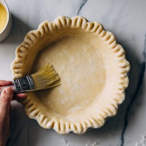 A hand brushing egg wash onto an unbaked pie crust set in a pie dish, with a cup containing a cracked egg nearby.