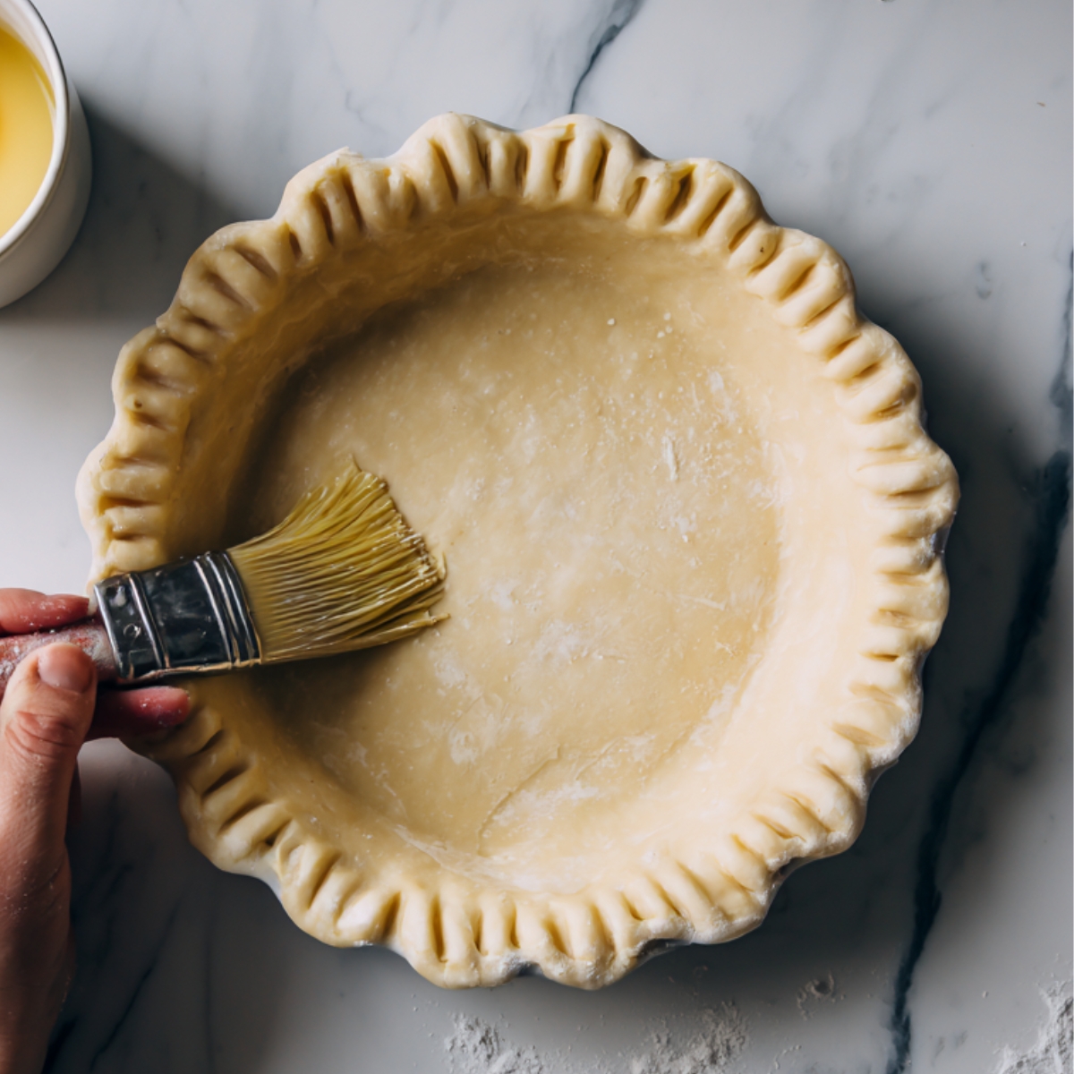 A hand brushing egg wash onto an unbaked pie crust set in a pie dish, with a cup containing a cracked egg nearby.