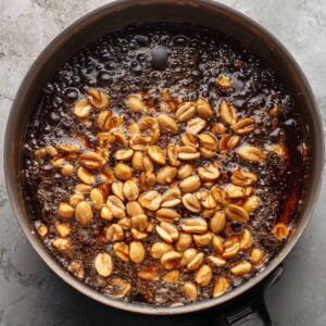 Step-by-step cooking shot of roasted peanuts being added into hot caramel mixture in a saucepan, overhead angle, food photography