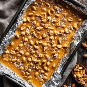 Overhead photo of freshly poured peanut brittle spread evenly in a foil-lined baking pan, shiny golden surface with peanuts, process step