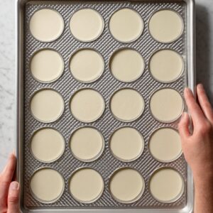 Overhead view of a baking sheet with evenly spaced unbaked macaron shells, hands holding the tray.