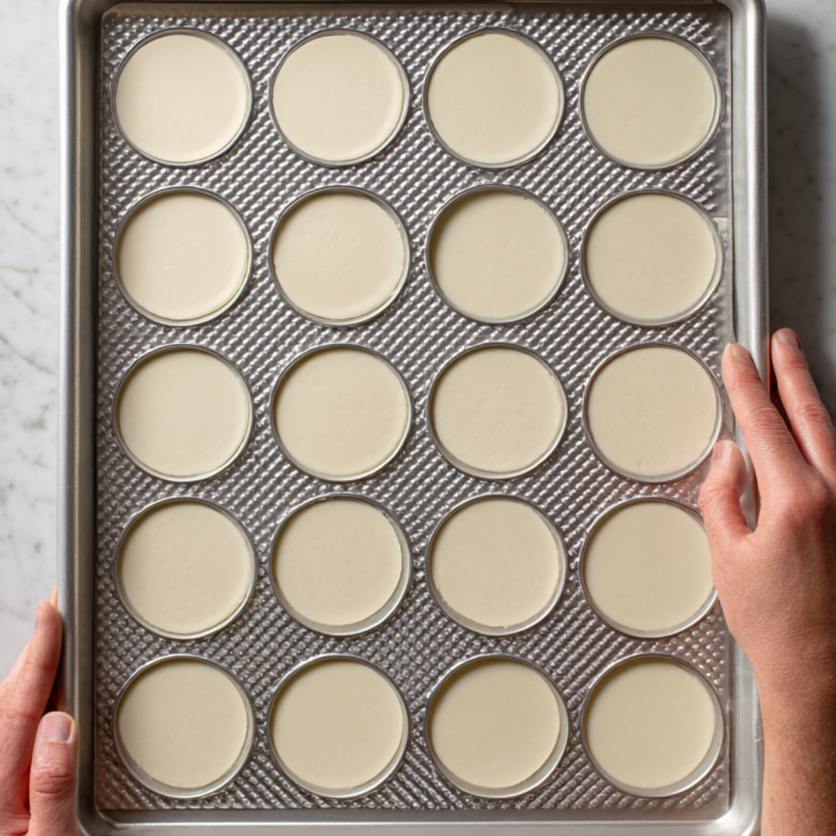 Overhead view of a baking sheet with evenly spaced unbaked macaron shells, hands holding the tray.