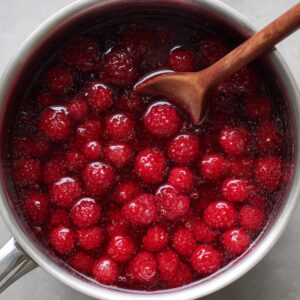 Pot of raspberries cooking with sugar, bubbling gently while being stirred with a wooden spoon.