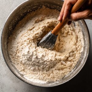 A hand mixing flour and dough in a metal bowl with a wooden spatula on a gray countertop.