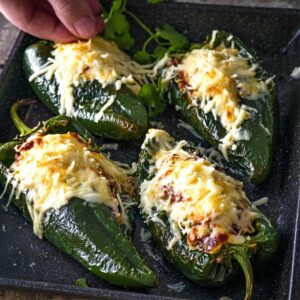 A baking tray with four stuffed poblano peppers topped with melted cheese, while a hand sprinkles fresh cilantro over them.