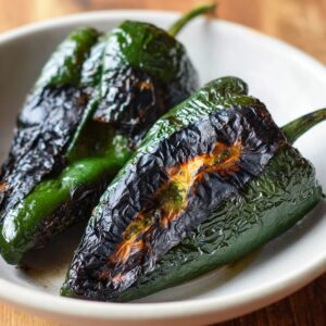 Two charred poblano peppers in a white bowl, blackened and blistered from roasting.