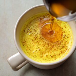 Golden turmeric milk in a mug with honey being poured on top, showing a swirl pattern and sprinkled spices