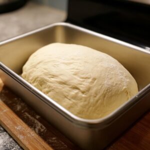 Loaf pan with risen bread dough placed on a wooden board, ready for baking.