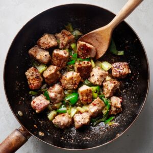 A wok filled with seared pork cubes: browned pork chunks cooked with chopped green peppers and onions, stirred with a wooden spoon.