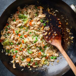 A black wok of fried rice in progress: white rice stir-fried with green peas, carrots, and bell peppers, plus chicken pieces. On the side, a pool of dark soy sauce with a wooden spatula resting.