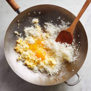 A silver wok of rice and eggs cooking together: cold white rice mixed with fresh cracked eggs, some scrambled and some still raw.
