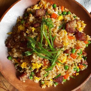 A large white bowl of finished pork fried rice: golden brown rice with scrambled eggs, carrots, broccoli, and pork chunks, topped with sliced green onions.