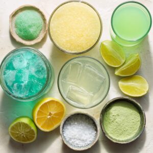 Several glasses and bowls with yellow, green, and blue drinks, along with cut lemons, limes, coarse salt, and green powder.