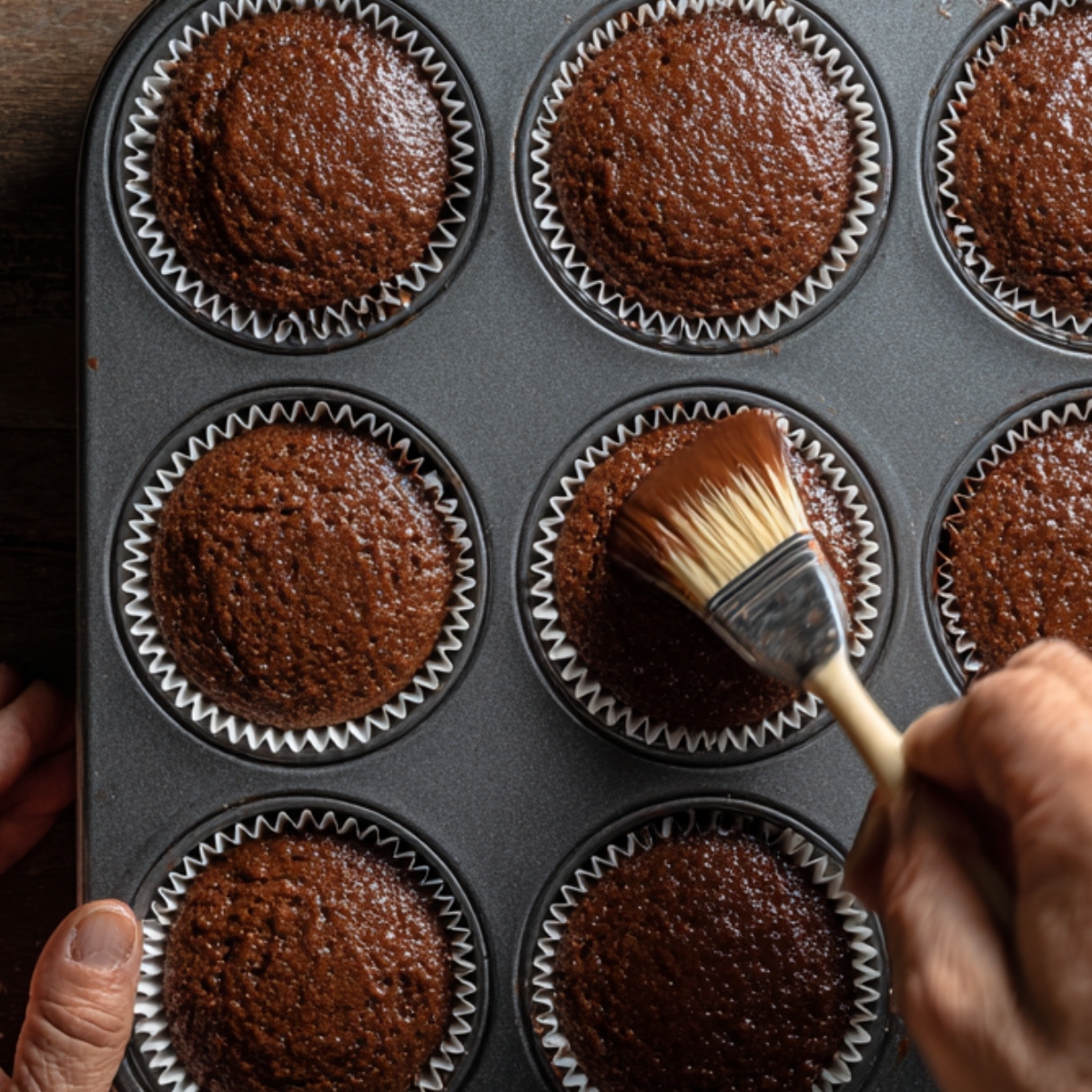 A muffin tin of freshly baked chocolate cupcakes, with a hand using a pastry brush to apply syrup or glaze.