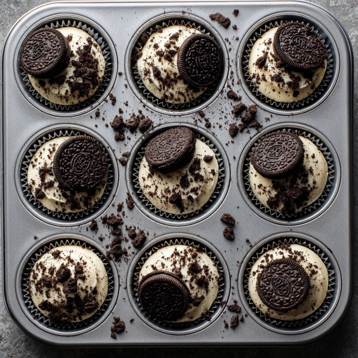 A cupcake tin filled with frosted cupcakes, each topped with an Oreo cookie and sprinkled with crumbs.