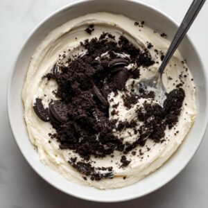 A white mixing bowl filled with creamy frosting, topped with crushed Oreo cookies, with a spoon resting inside.