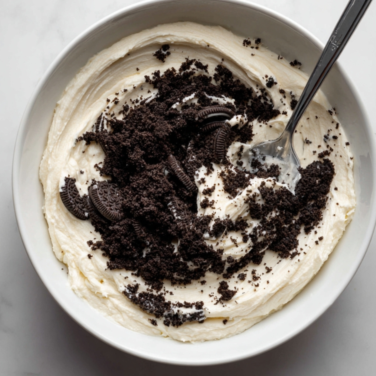 A white mixing bowl filled with creamy frosting, topped with crushed Oreo cookies, with a spoon resting inside.