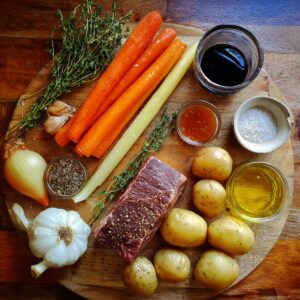 “The main pot roast ingredients displayed: chuck roast, carrots, potatoes, onions, garlic, celery, fresh thyme, olive oil, salt, pepper, broth, and seasonings.”