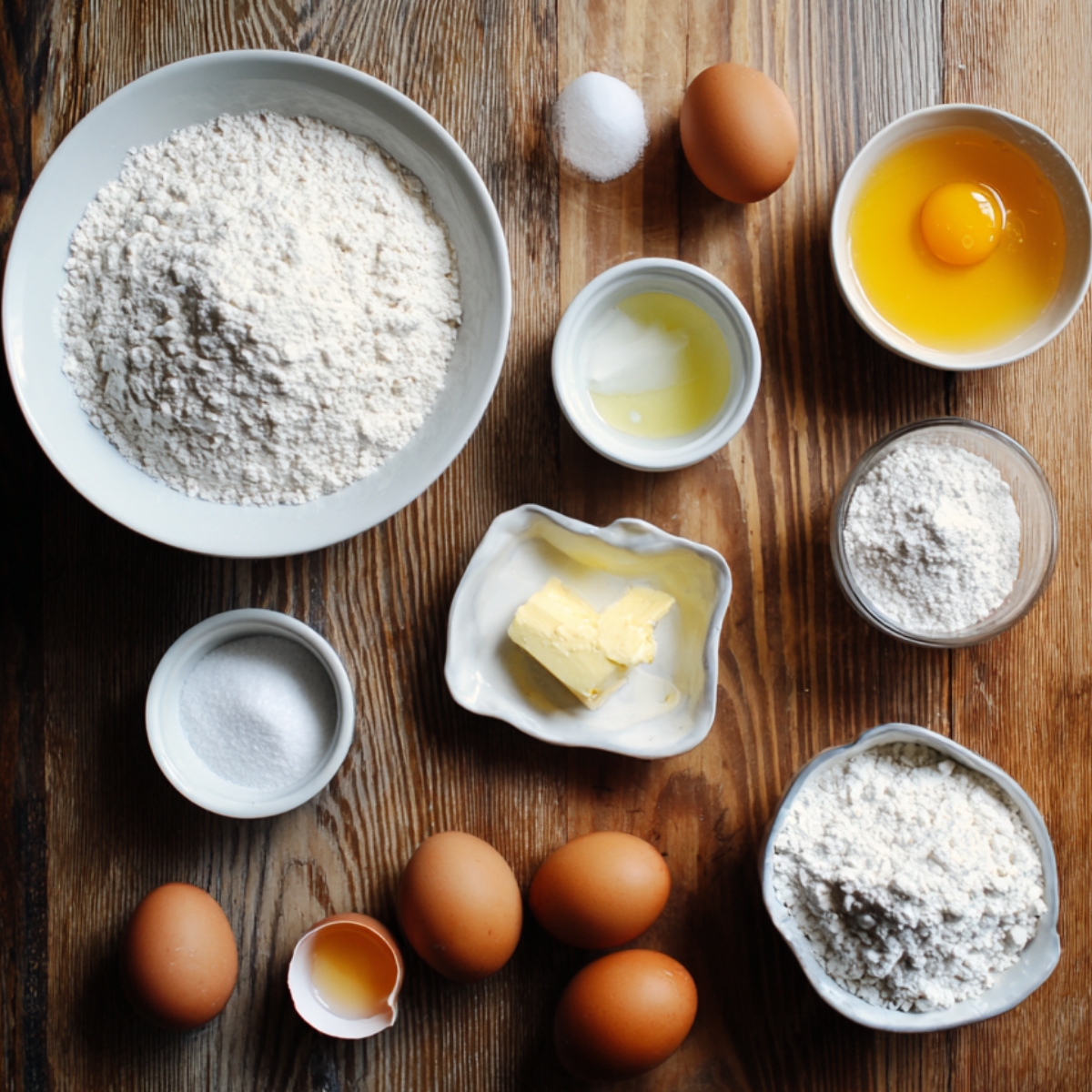 Several small bowls filled with flour, eggs, butter, sugar, and salt on a rustic wooden table.
