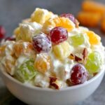 Small white bowl of creamy fruit Ambrosia Salad Recipe with red and green grapes, pineapple, orange, and nuts.