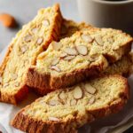 A small stack of Almond Biscotti Recipe on parchment paper, scattered whole almonds around, with a coffee cup in the background.