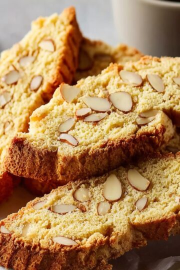 A small stack of Almond Biscotti Recipe on parchment paper, scattered whole almonds around, with a coffee cup in the background.