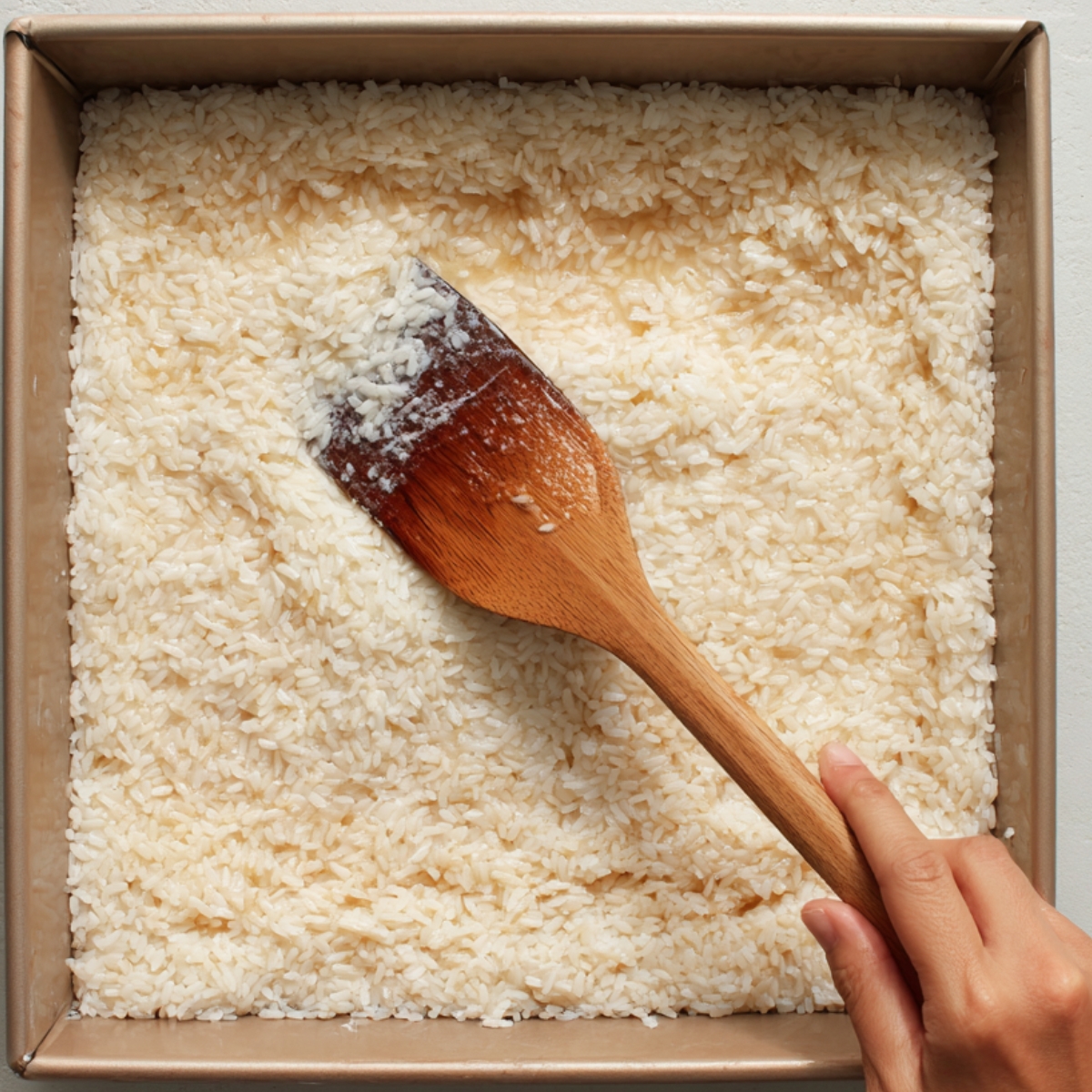 Cooked sticky rice being spread smoothly in a square baking pan using a wooden spatula, preparing the base layer for biko or another rice cake dessert.