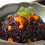 Cooked black rice recipe served in a green bowl, topped with diced orange vegetables, dried fruits, and a sprig of fresh herbs.