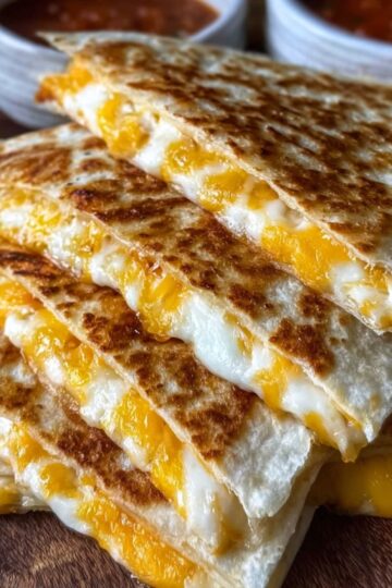 Sliced Cheese Quesadilla Recipe stacked on a plate, golden and cheesy, served with a bowl of red dipping sauce topped with herbs.
