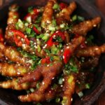 A wooden bowl filled with braised chicken feet recipe glazed in a glossy sauce, garnished with sliced red chili, chopped scallions, cilantro, and sprinkled sesame seeds.