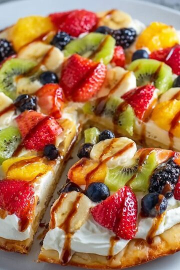A sliced dessert pizza recipe topped with whipped cream and a variety of fresh fruits like strawberries, bananas, kiwi, blueberries, and a drizzle of chocolate or caramel sauce