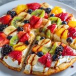 A sliced dessert pizza recipe topped with whipped cream and a variety of fresh fruits like strawberries, bananas, kiwi, blueberries, and a drizzle of chocolate or caramel sauce