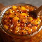 A glass jar of thick Mango Chutney Recipe, golden-orange in color, with visible mustard seeds and spices.