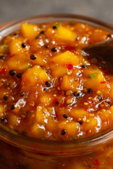 A glass jar of thick Mango Chutney Recipe, golden-orange in color, with visible mustard seeds and spices.