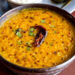 Masoor Dal Recipe A serving bowl of cooked dal garnished with fried red chili and cilantro, served with steamed rice, paratha, and pickle on the side.
