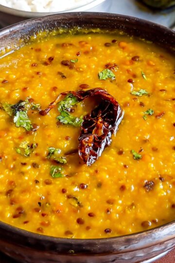 Masoor Dal Recipe A serving bowl of cooked dal garnished with fried red chili and cilantro, served with steamed rice, paratha, and pickle on the side.