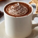 A white mug filled with rich Paris Hot Chocolate , topped with a swirl of whipped cream and dusted with cocoa, served on a saucer with croissants and a small Eiffel Tower model in the background.