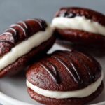 Finished Whoopie Pie Recipe: Chocolate whoopie pies filled with white cream and drizzled with chocolate, served on a small white plate.