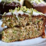 A moist slice of Zucchini Cake Recipe topped with white frosting and sprinkled with chopped pistachios, served on a ceramic plate.