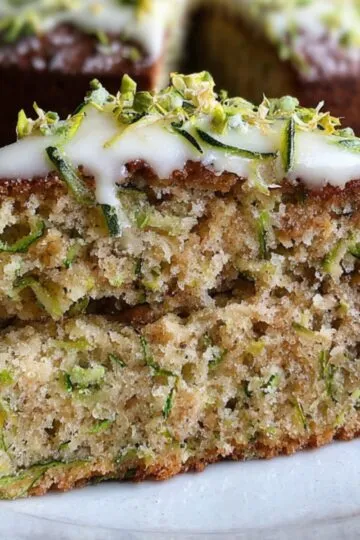 A moist slice of Zucchini Cake Recipe topped with white frosting and sprinkled with chopped pistachios, served on a ceramic plate.