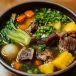Another serving of bulalo recipe Filipino food — A black bowl containing beef shank, corn, carrots, potatoes, and bok choy in broth, garnished generously with chopped herbs and scallions.