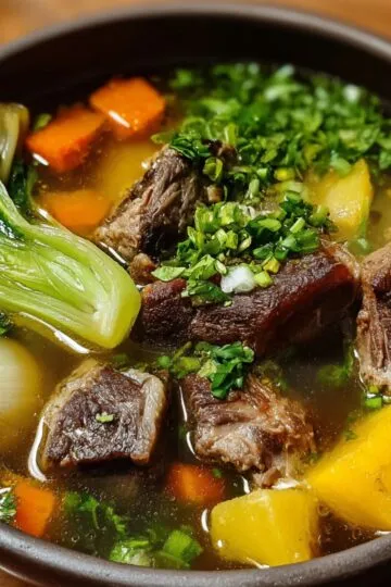 Another serving of bulalo recipe Filipino food — A black bowl containing beef shank, corn, carrots, potatoes, and bok choy in broth, garnished generously with chopped herbs and scallions.