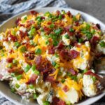 A serving bowl of loaded baked potato salad with shredded cheese, crispy bacon, and chopped green onions.