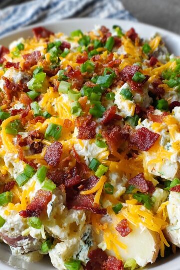 A serving bowl of loaded baked potato salad with shredded cheese, crispy bacon, and chopped green onions.
