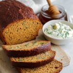 A loaf of rustic homemade outback bread recipe, partially sliced, served with a bowl of creamy herb butter and honey in the background.